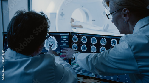 Canvas Print In Control Room Doctor and Radiologist Discuss Diagnosis while Watching Procedure and Monitors Showing Brain Scans Results, In the Background Patient Undergoes MRI or CT Scan Procedure