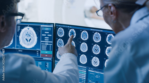 Canvas Print In Control Room Doctor and Radiologist Discuss Diagnosis while Watching Procedure and Monitors Showing Brain Scans Results, In the Background Patient Undergoes MRI or CT Scan Procedure