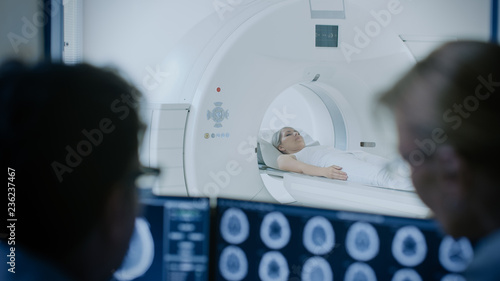 In Control Room Doctor and Radiologist Discuss Diagnosis while Watching Procedure and Monitors Showing Brain Scans Results, In the Background Patient Undergoes MRI or CT Scan Procedure.