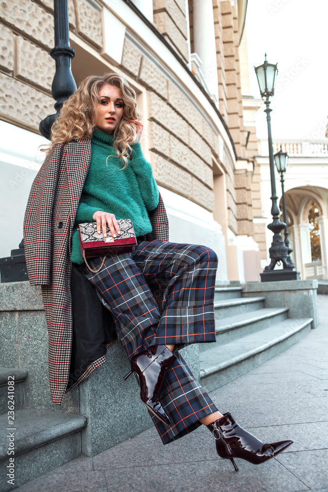 Outdoor full body fashion portrait of young beautiful woman wearing stylish green sweater, checkered pants, trendy sock boots, holding small purple bag, posing in street. Copy space.