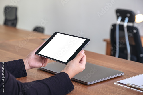Mockup image of hands holding and using black tablet pc with blank white desktop screen with notebook on wooden table in office