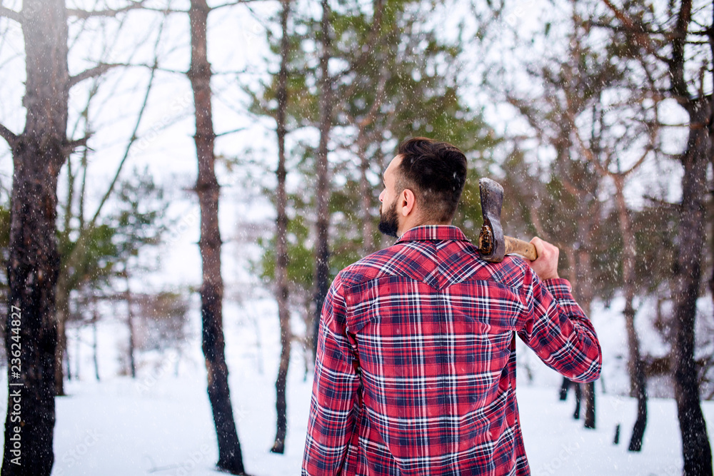 Bearded hipster man in a winter snowy forest with axe on a shoulder. Woodman standing in the forest. Male inspecting trees in woods. Lumberjack woodcutter holding ax wearing plaid red shirt.