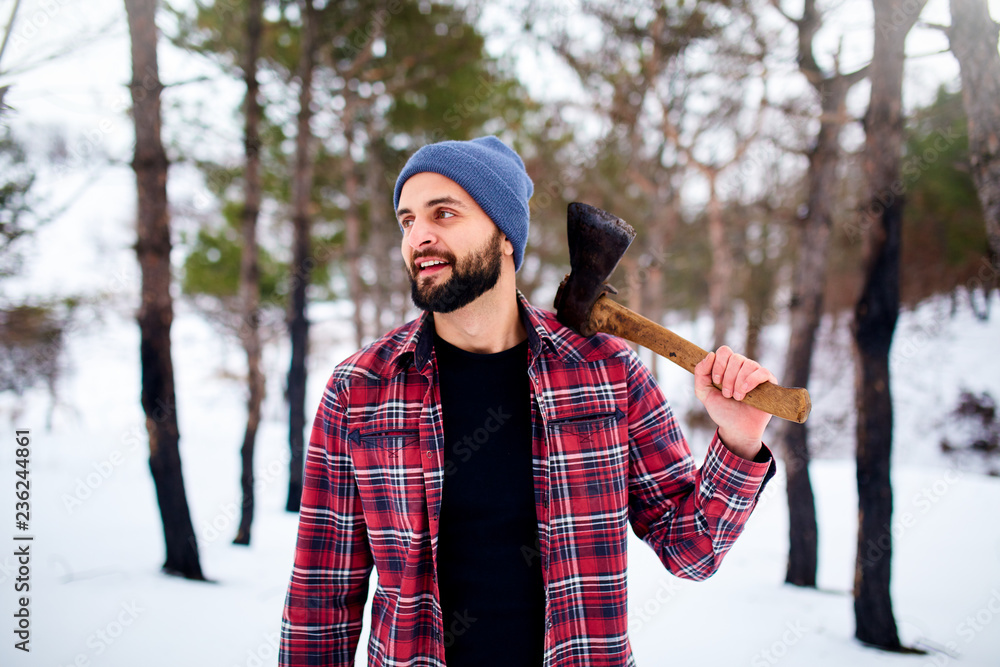 Fotka „Bearded hipster man in a winter snowy forest with axe on a shoulder.  Woodman standing in the forest. Male inspecting trees in woods. Lumberjack  woodcutter holding ax wearing plaid red shirt.“ ze