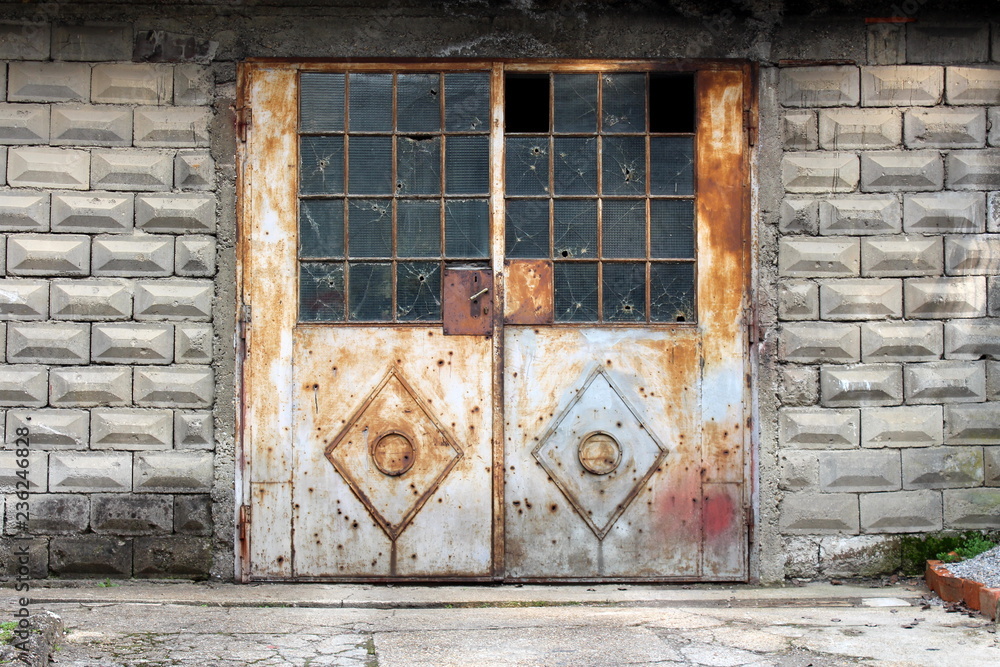 Grey partially rusted metal doors with small door handle and broken safety wire protection glass mounted on concrete wall with stone tiles with concrete driveway in front Grey partially rusted metal doors with small door handle and broken safety wire protection glass mounted on concrete wall with stone tiles with concrete driveway in front