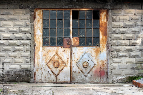 Grey partially rusted metal doors with small door handle and broken safety wire protection glass mounted on concrete wall with stone tiles with concrete driveway in front