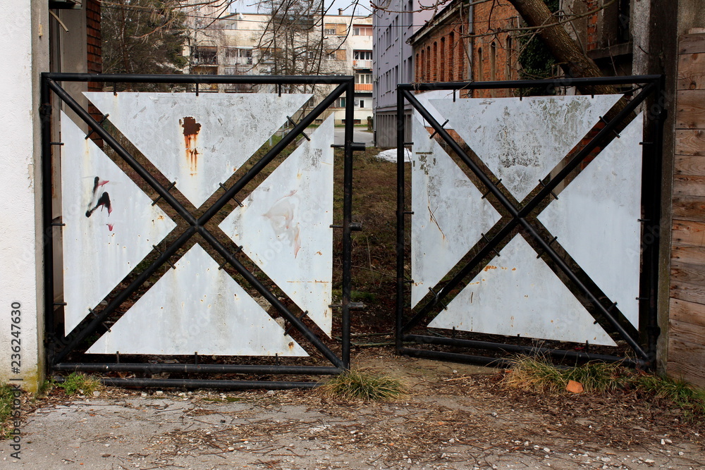 Partially rusted open heavy metal doors with black frame and white to grey sheet metal fillings preventing access to abandoned backyard Partially rusted open heavy metal doors with black frame and white to grey sheet metal fillings preventing access to abandoned backyard