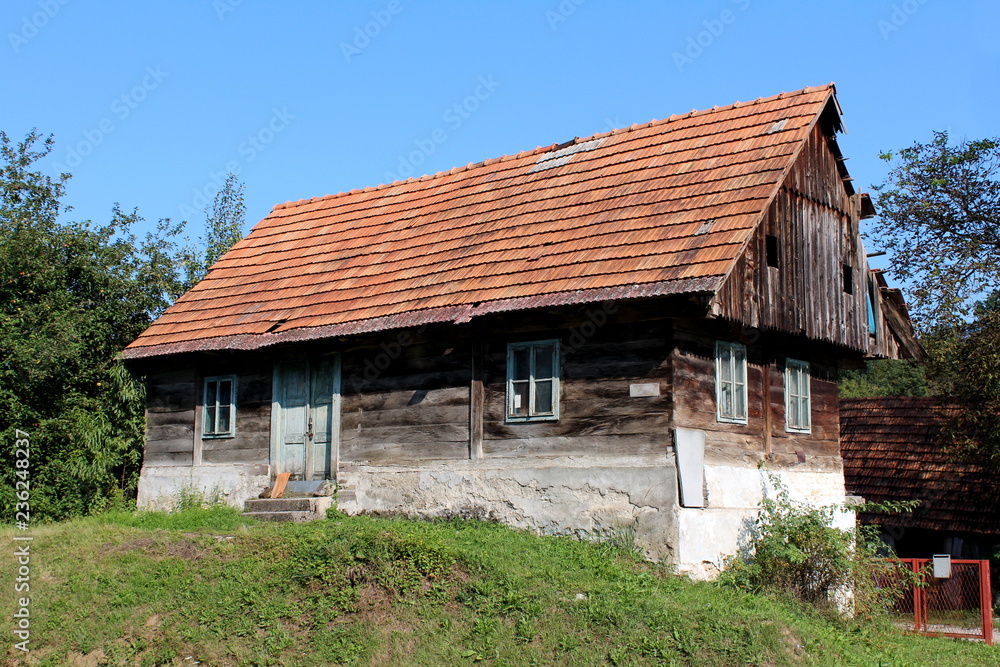 Small wooden old family house with dilapidated boards and broken roof with missing roof tiles on top of small hill surrounded with grass and trees with clear blue sky in background