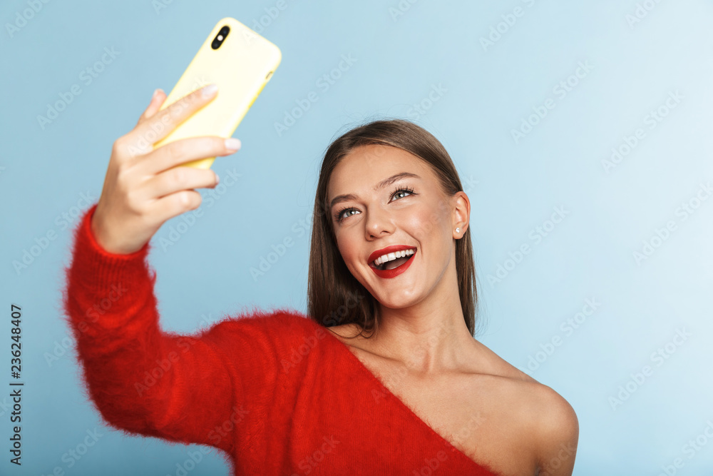 Emotional excited young woman posing isolated over blue wall background take a selfie by mobile phone. Emotional excited young woman posing isolated over blue wall background take a selfie by mobile phone.