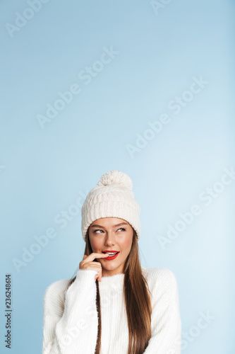 Cute thinking young woman wearing winter hat posing isolated over blue wall background.