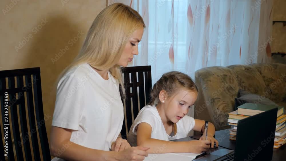Beautiful woman with little diligent girl sitting at table in living room having textbooks and laptop while doing homework Beautiful woman with little diligent girl sitting at table in living room having textbooks and laptop while doing homework