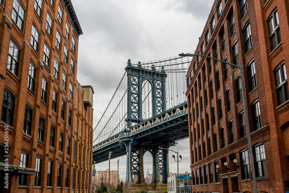 Fototapeta premium Manhattan Bridge Oglądany Z Dumbo, Brooklyn, Nowy Jork między dwoma budynkami z czerwonej cegły i pochmurne tło