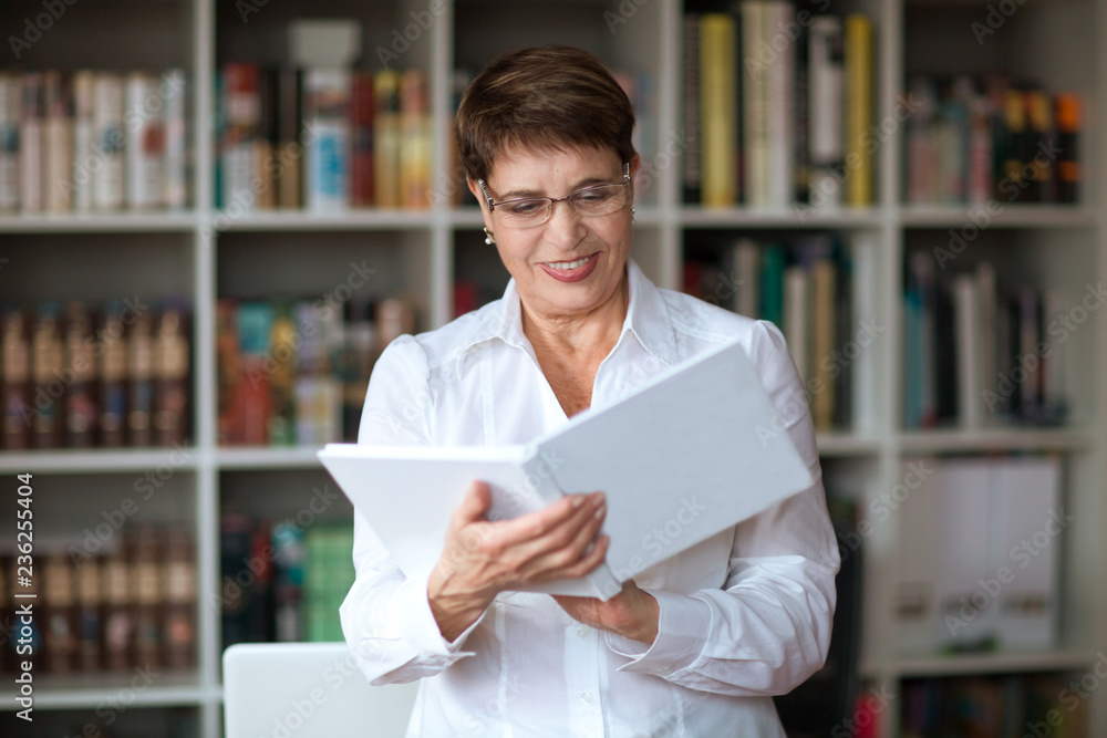 Happy senior woman wearing glasses head shot in a white shirt,  with book against the background of a bookcase