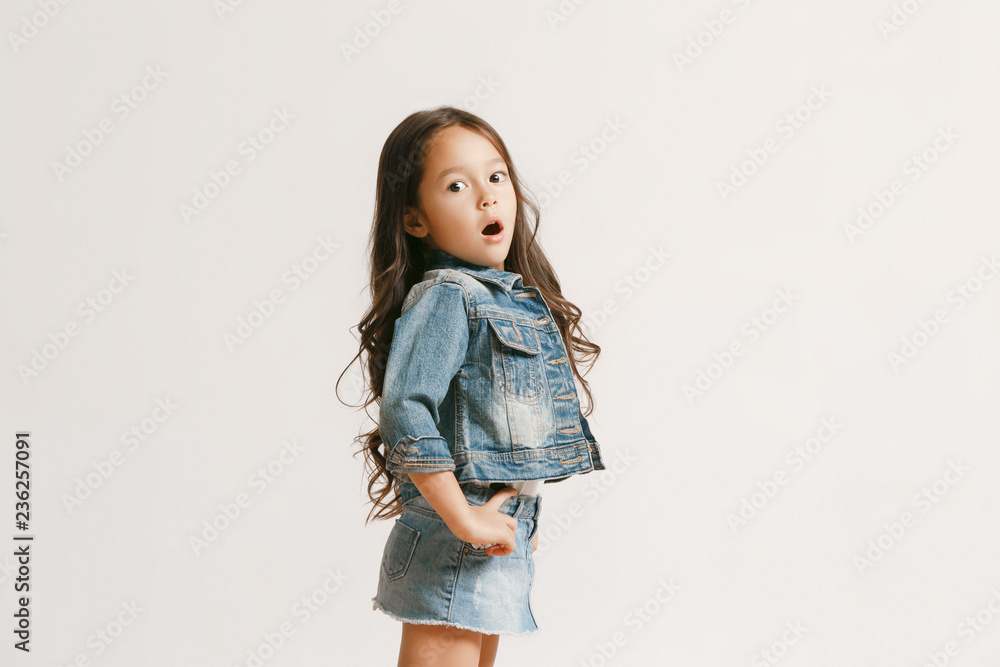 The portrait of cute little kid girl in stylish jeans clothes looking at  camera and smiling, standing against white studio wall. Kids fashion  concept Stock Photo