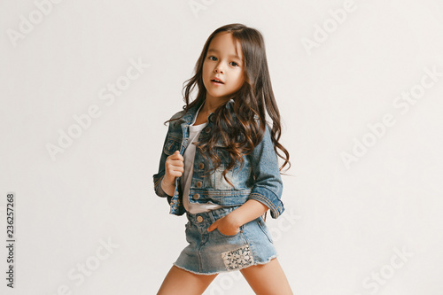 Canvastavla Full length portrait of cute little kid girl in stylish jeans clothes looking at camera and smiling, standing against white studio wall