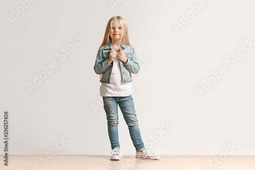 Full length portrait of cute little kid girl in stylish jeans clothes looking at camera and smiling, standing against white studio wall. Kids fashion concept