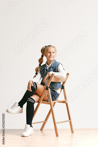 Full length portrait of cute little teen girl in stylish jeans clothes looking at camera and smiling against white studio wall. Kids fashion concept