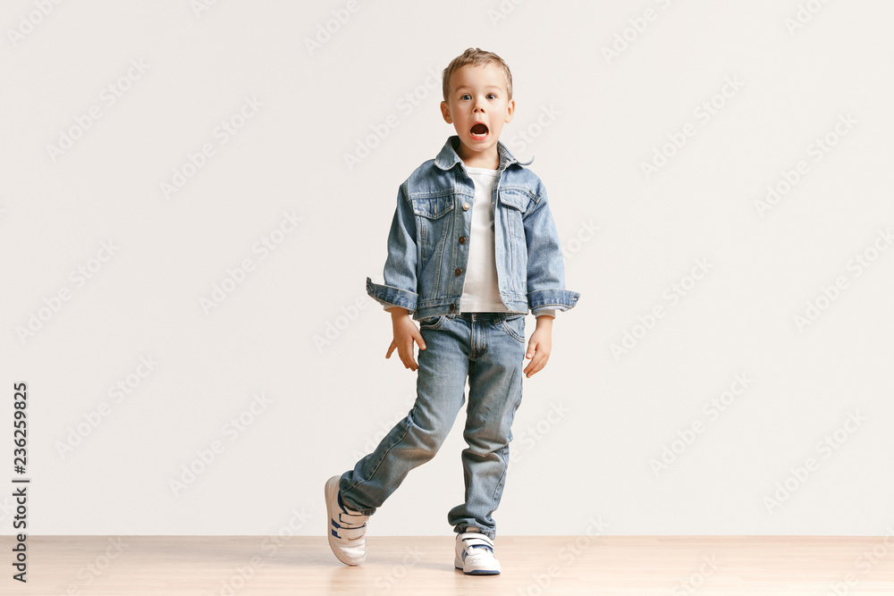 The portrait of cute little kid boy in stylish jeans clothes looking at camera against white studio wall. Kids fashion concept