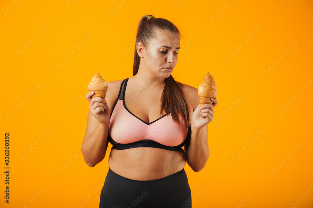 Image of serious chubby woman in tracksuit holding two croissants in both hands, isolated over yellow background Image of serious chubby woman in tracksuit holding two croissants in both hands, isolated over yellow background