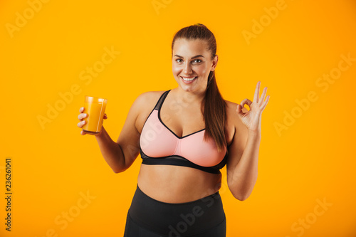 Image of healthy chubby woman in tracksuit smiling and holding glass with orange juice, isolated over yellow background