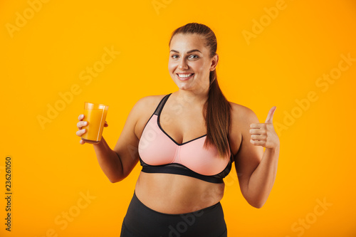 Image of beautiful chubby woman in tracksuit smiling and holding glass with orange juice, isolated over yellow background
