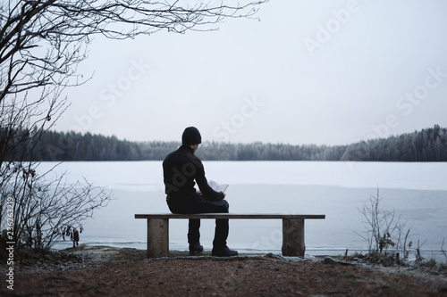 A man is reading a book on a bench on the bank of a frozen lake. Top branches hang down. The foreground is in focus, the horizon is blurred.