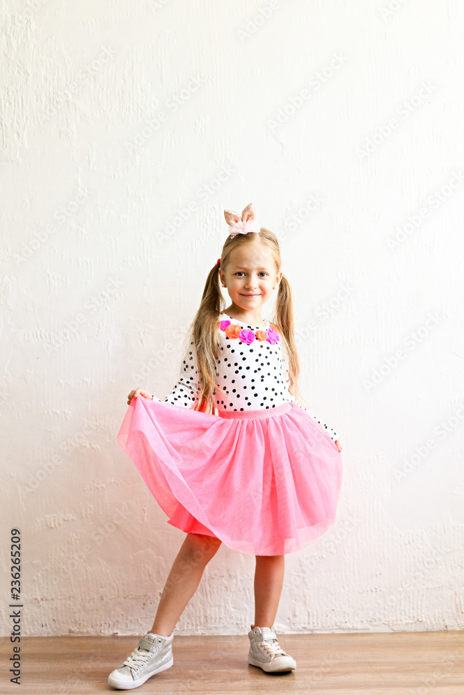 Portrait of cute little five year old girl with long blond hair wearing easter bunny ears on head, colorful polka dot shirt and pink tutu skirt. White wall backgroung, copy space, close up.