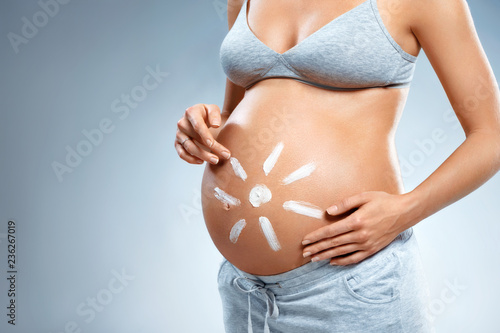 Close up of pregnant woman applying moisturizing cream on her belly on grey background. Pregnancy, maternity, preparation and expectation concept