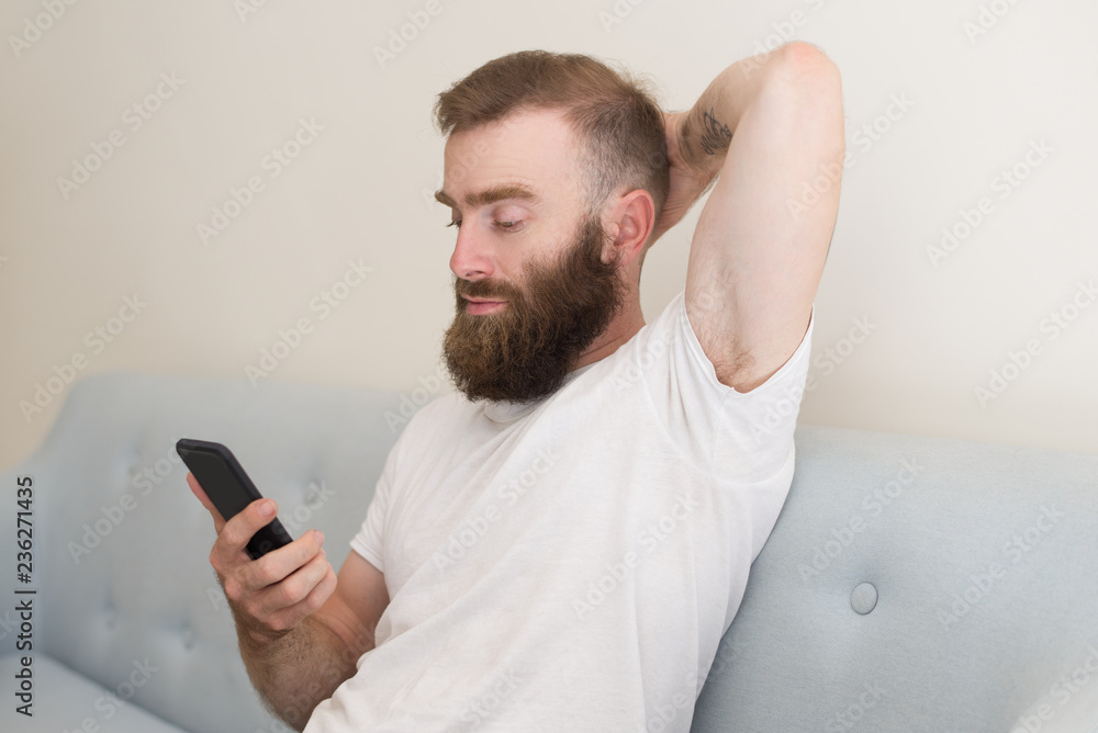 Focused man browsing on smartphone and sitting on sofa. Handsome guy keeping hand behind head and relaxing. Communication concept. Front view. Focused man browsing on smartphone and sitting on sofa. Handsome guy keeping hand behind head and relaxing. Communication concept. Front view.