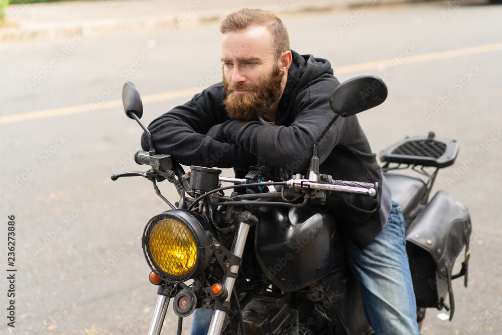 Portrait of man sitting on motorbike with pensive expression. Young Caucasian biker resting on his bike outdoors. Transportation concept