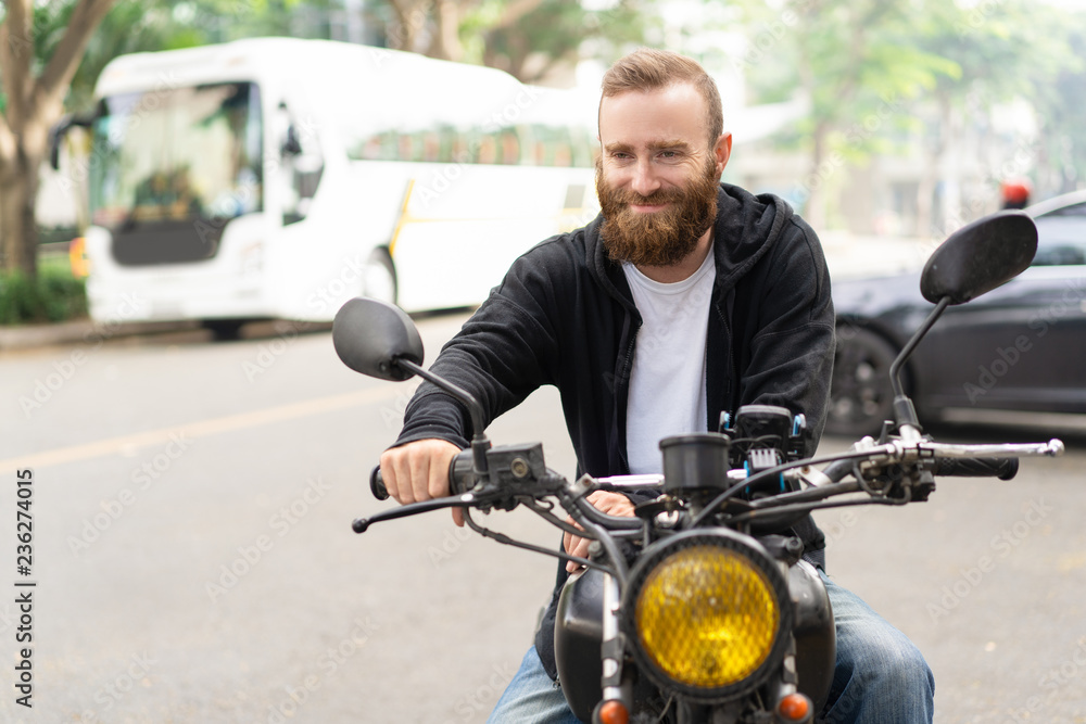 Fototapeta premium Portret uśmiechnięty rowerzysta siedzi na motocyklu na poboczu drogi. Młody mężczyzna kaukaski jedzie motocykl w mieście. Koncepcja kultury rowerzysty