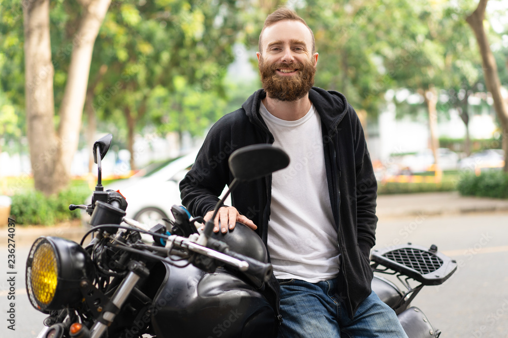 Portrait of smiling young man sitting on his motorcycle. Happy biker posing with motorbike outdoors. Biker lifestyle concept