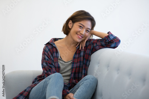 Cheerful Asian girl in casual enjoying weekend. Beautiful young woman in checkered shirt and jean resting on couch and smiling at camera. Weekend and leisure concept