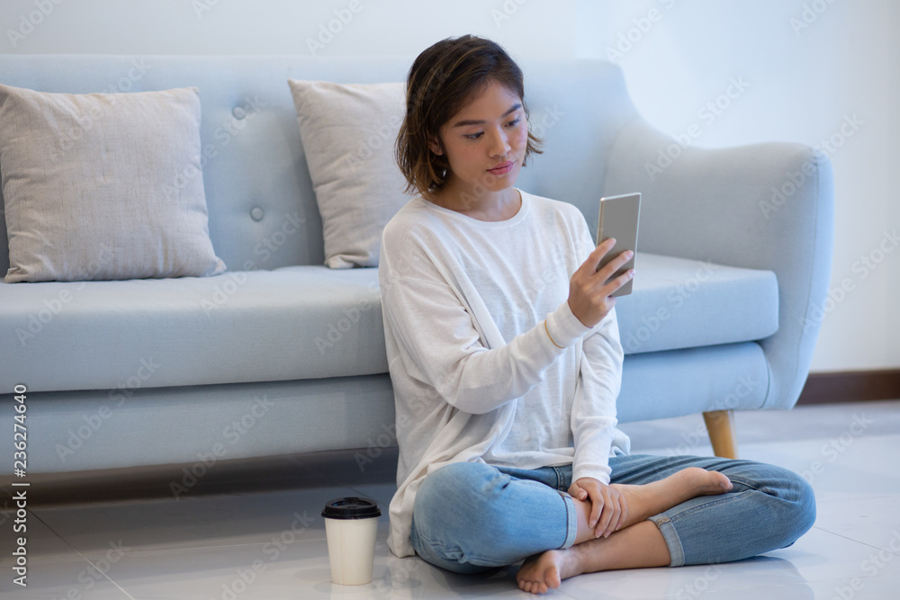 Focused Asian girl taking selfie at home. Beautiful young woman sitting in front of couch in lotus position and photographing by smartphone. Photography concept Focused Asian girl taking selfie at home. Beautiful young woman sitting in front of couch in lotus position and photographing by smartphone. Photography concept