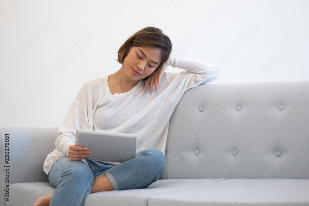 Pensive dreamy Asian girl enjoying weekend at home and watching movie on tablet. Young woman in casual sitting on couch and looking at screen. Connection concept Pensive dreamy Asian girl enjoying weekend at home and watching movie on tablet. Young woman in casual sitting on couch and looking at screen. Connection concept