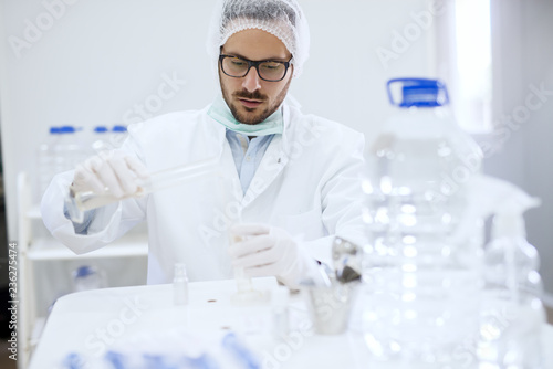 Scientist checking water quality at water factory. In front of him gallons with water. In hands test tubes. Protective suit on.