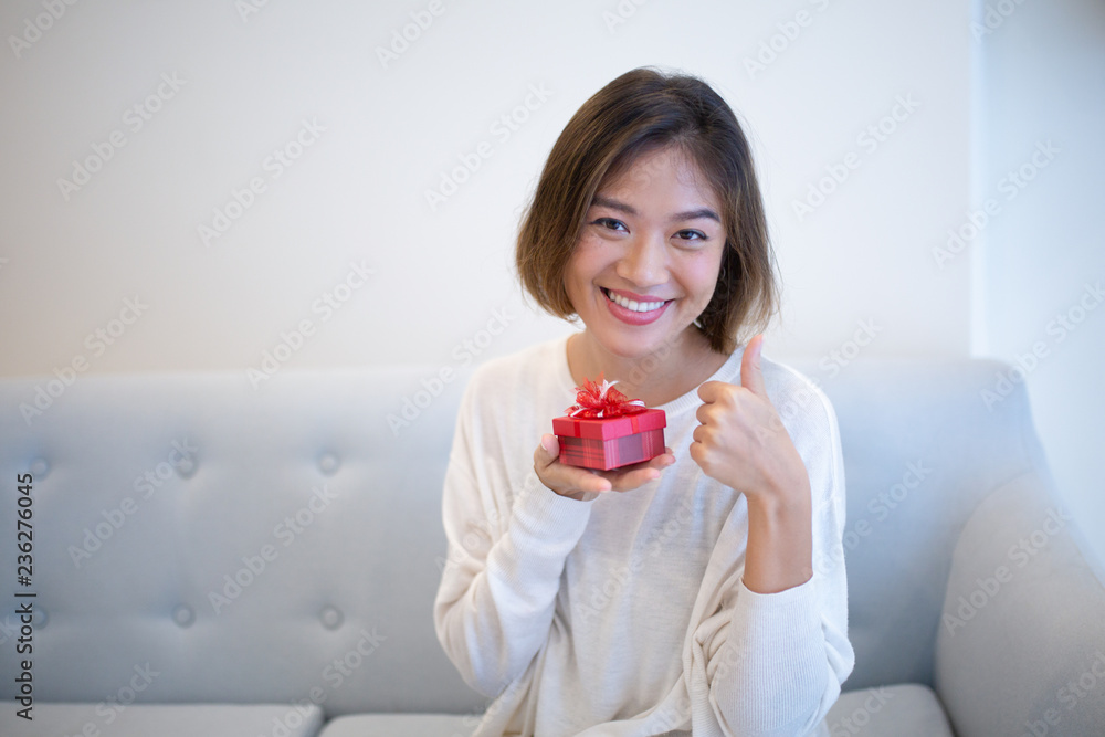 Positive excited woman with small gift box showing thumb-up and looking at camera. Jolly pretty young Asian lady in casual sweater sitting on elegant sofa. Christmas gift concept Positive excited woman with small gift box showing thumb-up and looking at camera. Jolly pretty young Asian lady in casual sweater sitting on elegant sofa. Christmas gift concept