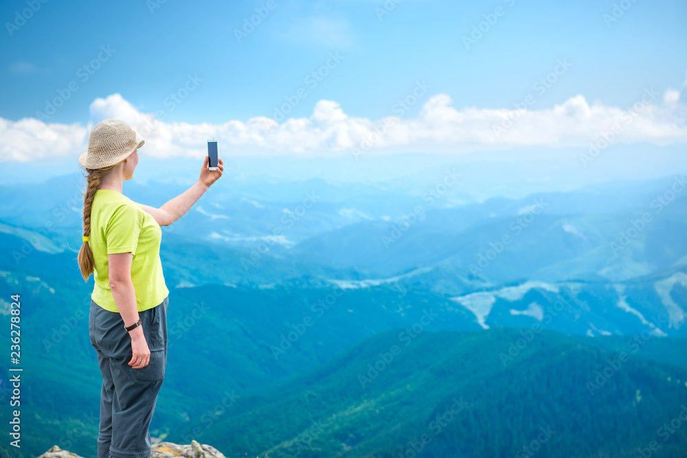 Young woman standing on the mountains cliff and taking travel selfie on smartphone