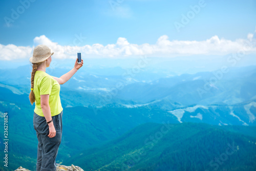 Young woman standing on the mountains cliff and taking travel selfie on smartphone
