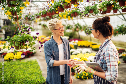 Фотография Female florist dressed in apron with tablet in hands shaking hands with customer while standing in greenhouse