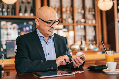 Bearded senior adult in suit using smart phone for reading or writing message while sitting in cafeteria. On desk credit tablet, coffee, wallet, and juice.