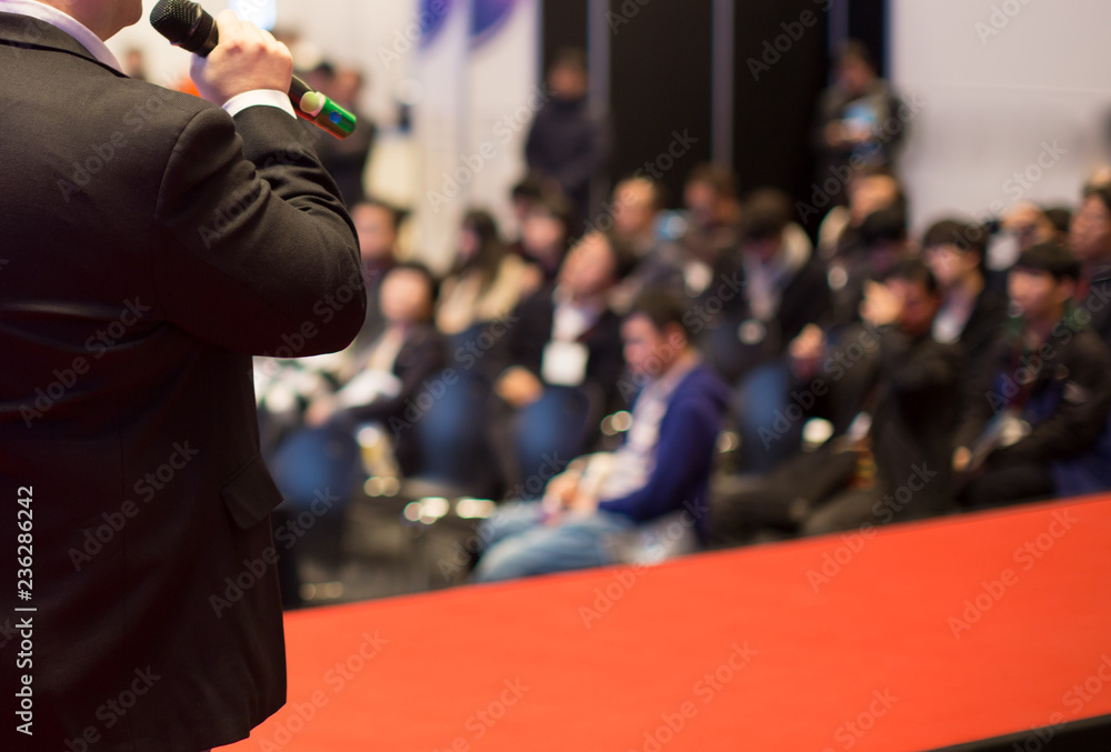 Plakat Businessman Presenter Giving a Speech to Group of People. Speaker on Stage Holding Microphone During Investor Pitch Presentation. Business Expert Manager Gives Talk at Conference with Audience and Red