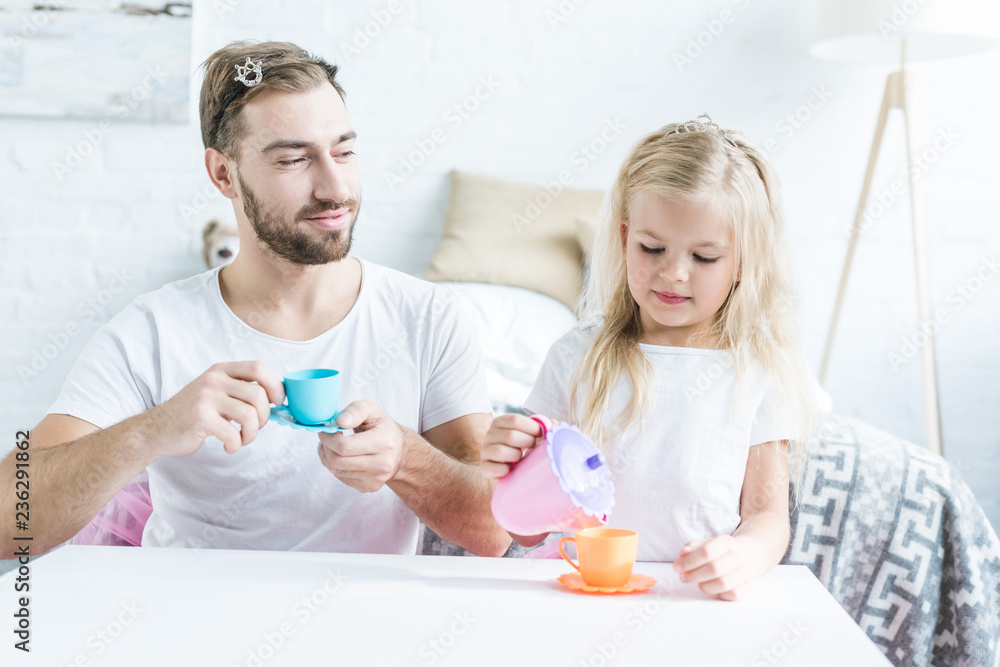 smiling father holding toy cup and looking at little daughter pouring tea from toy kettle smiling father holding toy cup and looking at little daughter pouring tea from toy kettle