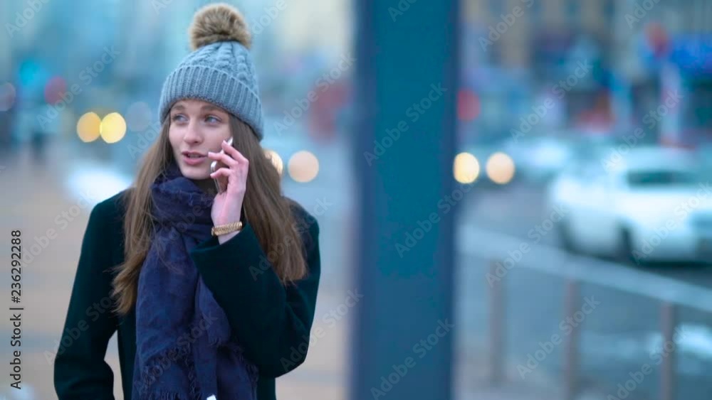 A young girl walking down the street and talking on the phone. Near ride cars. Winter and cold. Beautiful hat. A young girl walking down the street and talking on the phone. Near ride cars. Winter and cold. Beautiful hat.