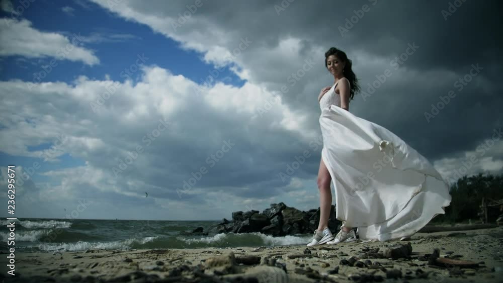 Beautiful girl in a white dress stands on seashore. Dress flutters in wind slow motion. 