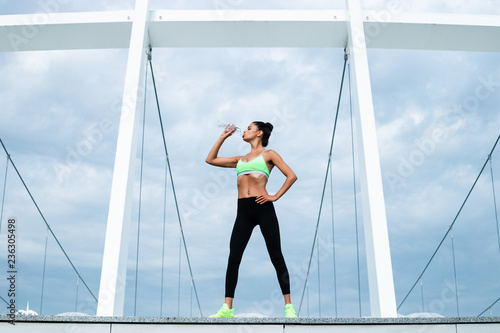 Water balance in her body. Full length of young beautiful fit woman in sportswear drinking water while standing against industrial city view