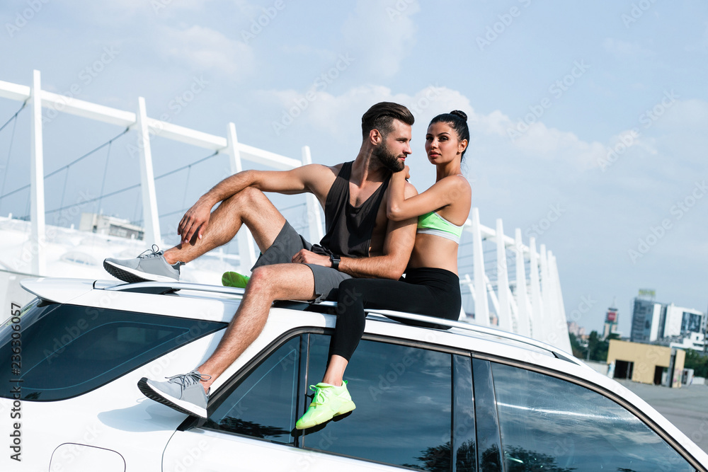 Their urban life. Young beautiful couple in sportswear bonding to each other while sitting on the car against industrial city view