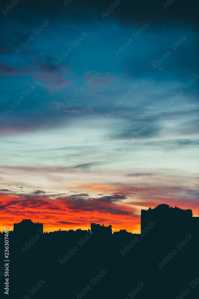 Cityscape with wonderful varicolored vivid dawn. Amazing dramatic multicolored cloudy sky above dark silhouettes of city buildings. Atmospheric background of sunrise in overcast weather. Copy space.