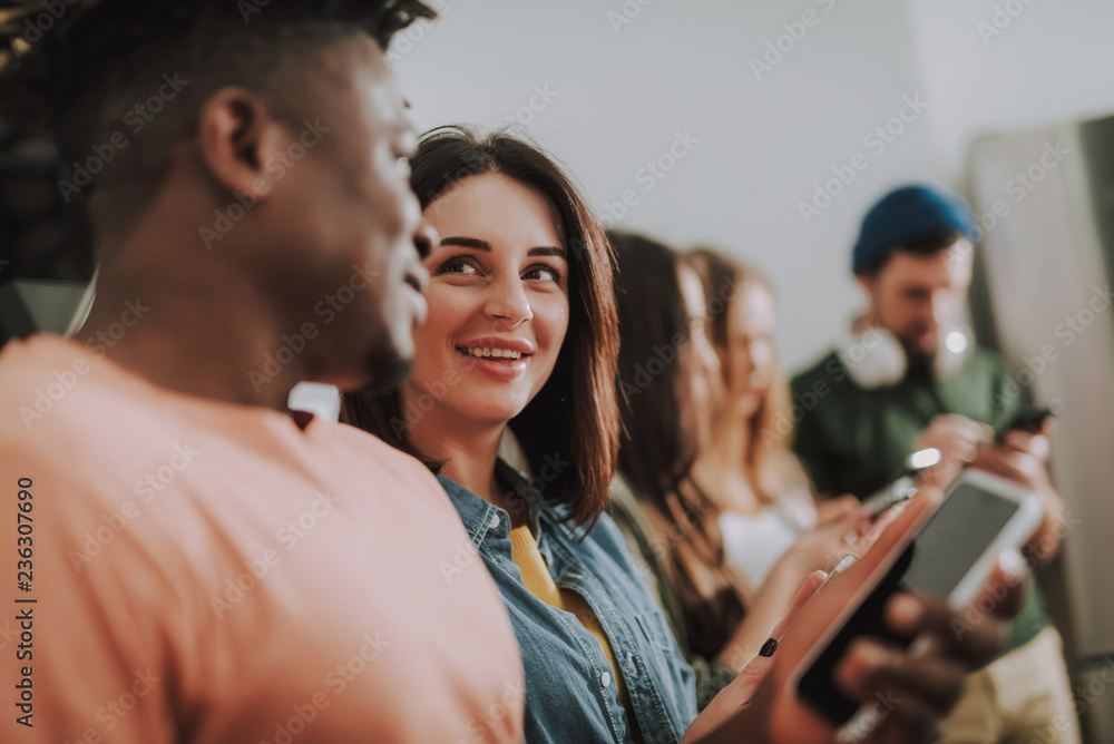 Obraz premium Portrait of beautiful girl gazing at friend and smiling while he holding smartphone. Bearded gentleman and two women on blurred background