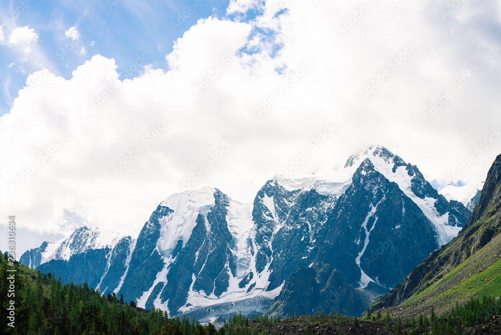 Snowy mountain top between rocky mountains under overcast sky. Rocky ridge in mist above forest. Atmospheric minimalistic landscape of majestic nature.