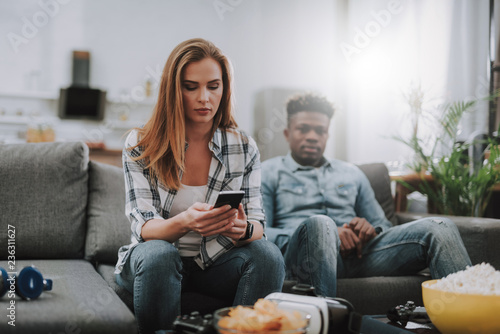 Portrait of charming girl holding smartphone and looking at it with serious expression. Handsome afro american guy sitting on couch on blurred background
